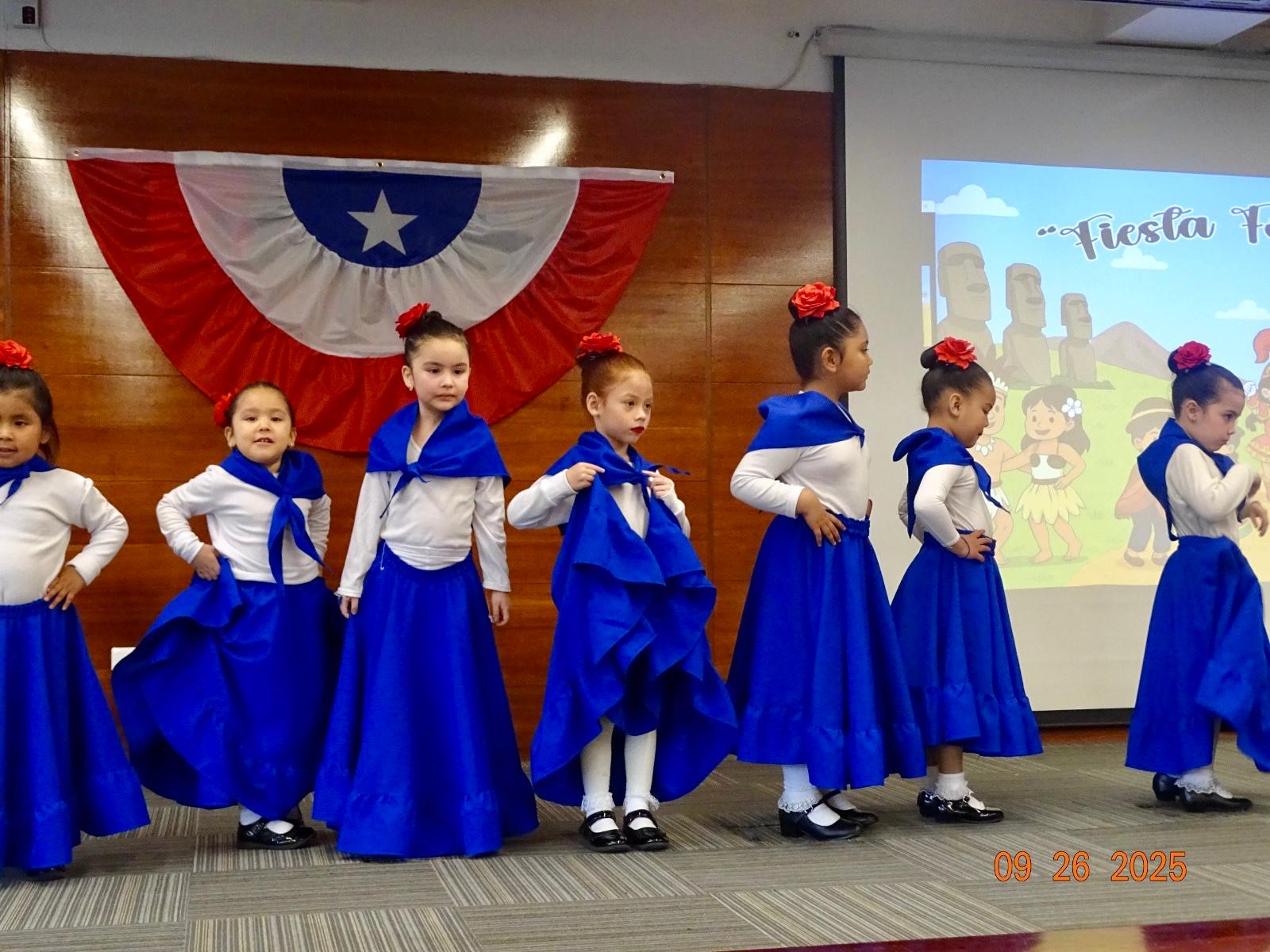 Niñas bailando cueca con faldas azules frente a la bandera chilena